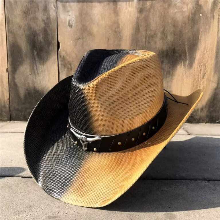 Two-tone black and golden brown straw cowboy hat with silver longhorn embellished band, featuring pinch front crown and curved brim, displayed against weathered wall